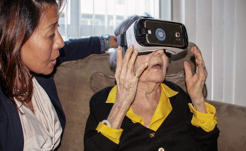 Two female women, one an older adult wearing virtual reality glasses and the other younger assisting her.